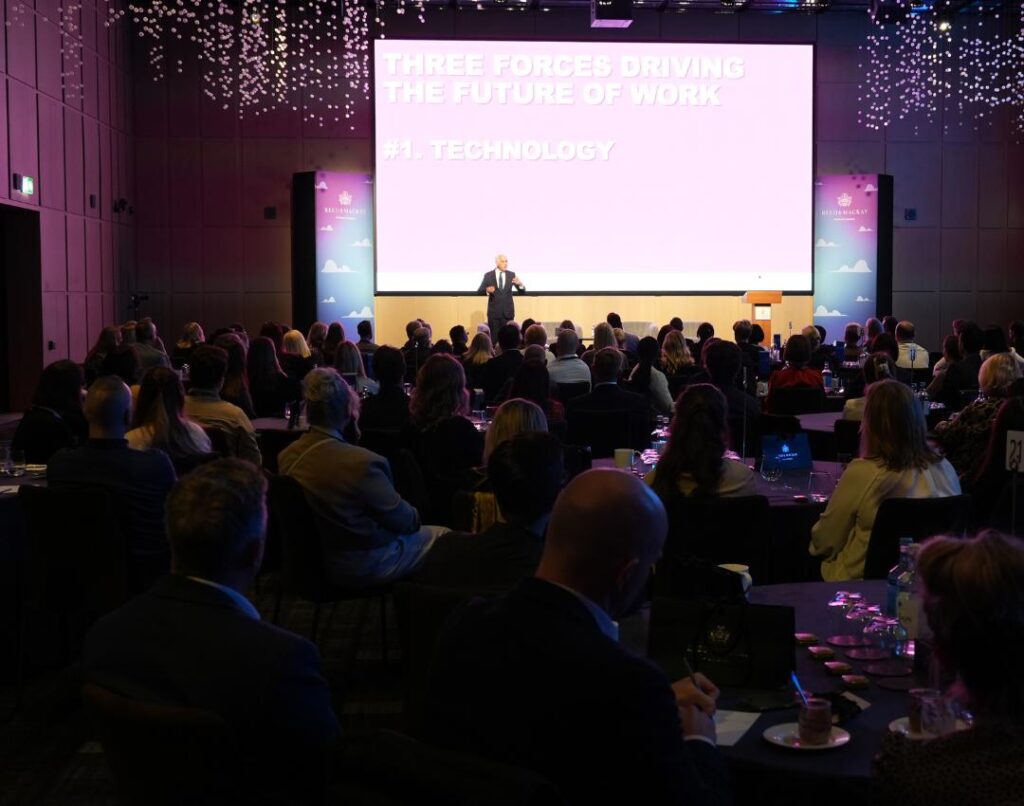 Speaker on stage speaking at a conference with the audience in front of them.