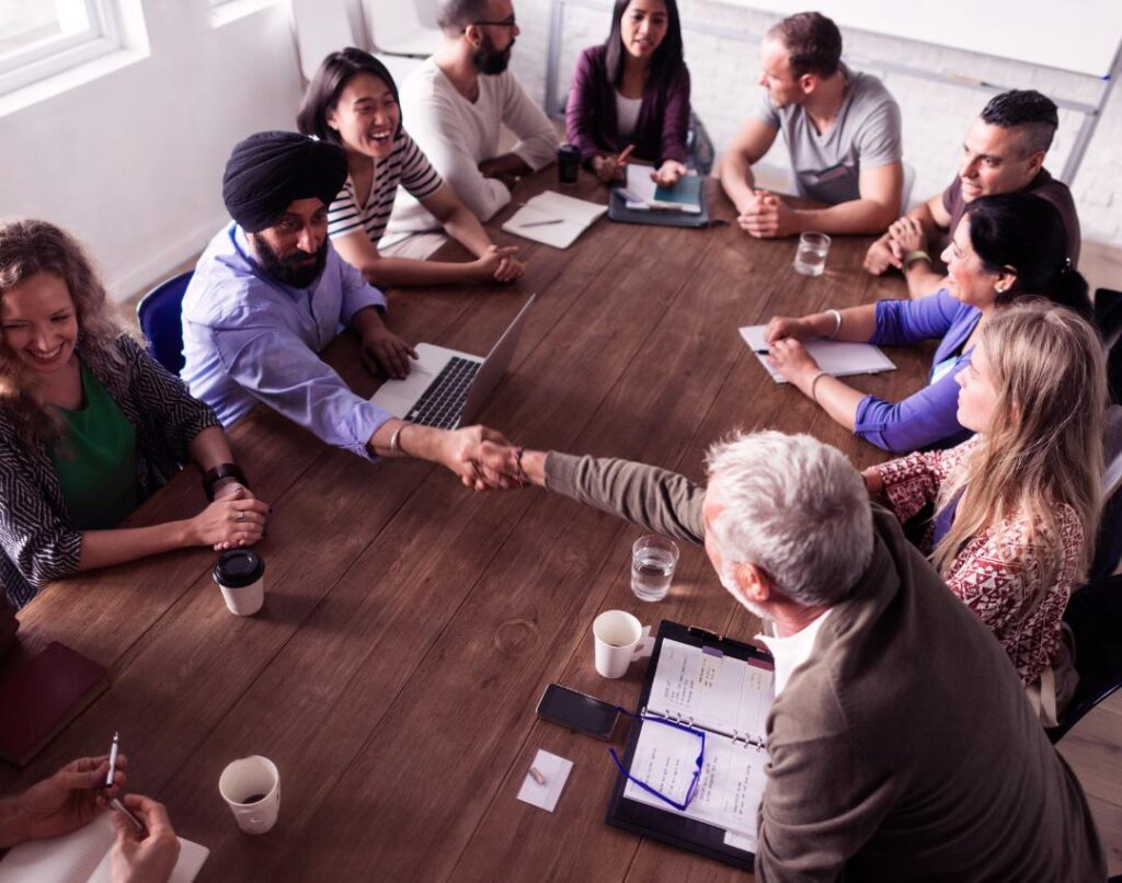 Group of employees sat around a table shaking hands. The employees are a range of ages.